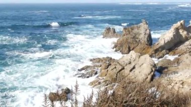 Kayalık okyanus plajı. Büyük dalgalar kayalıklara çarpıyor, mavi su sıçrıyor, deniz köpüğü. Big Sur yakınlarında doğanın gücü, 17 millik yol. Dramatik deniz manzarası. Point Lobos, Monterey, California sahili, ABD