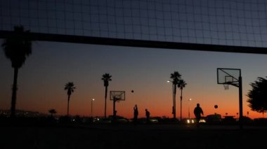 Basketbol sahasındaki oyuncuların siluetleri, basketbol oynayan insanlar, günbatımı okyanus plajı, Kaliforniya sahili, ABD. Sokak sporu sahasında siyah halka, ağ ve arkabahta. Görev plajı.
