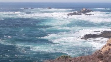 Kayalık okyanus plajı. Büyük dalgalar kayalıklara çarpıyor, mavi su sıçrıyor, deniz köpüğü. Big Sur yakınlarında doğanın gücü, 17 millik yol. Dramatik deniz manzarası. Point Lobos, Monterey, California sahili, ABD