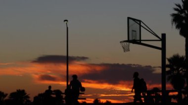 Basketbol oynayan insanlar, basketbol sahasındaki oyuncuların siluetleri, günbatımı okyanus sahili, California sahili, Mission plajı, ABD. Sokak topu sahasında siyah halka, ağ ve arkabahta.
