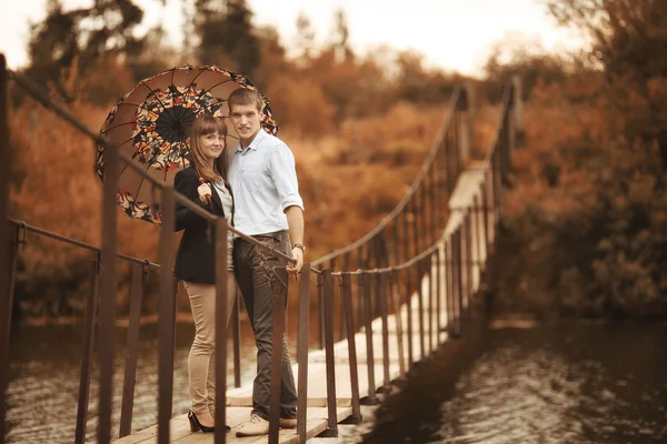 Young lovers on a rope bridge across the river under the umbrella — Stock Photo, Image