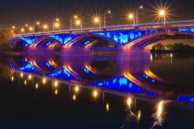 Toplumsal köprüsü olan karanlık şehir manzarası ve Rusya 'nın Krasnoyarsk kentindeki Yenisei Nehri' nin sularında renkli bir aydınlanmanın yansıması. Gece şehir ışıkları