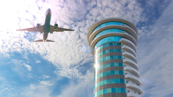Low-flying passenger commercial plane above glass air traffic control tower against blue sky with clouds backgrounds. Vacation holiday travel. Air transportation background with copy space