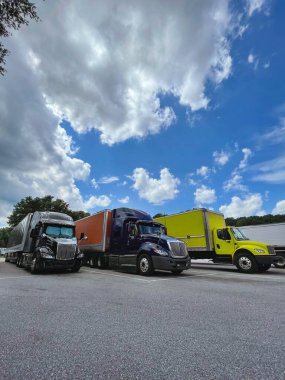 Three large, colorful trucks parked on asphalt at a highway rest stop under a bright blue sky with dramatic clouds. 