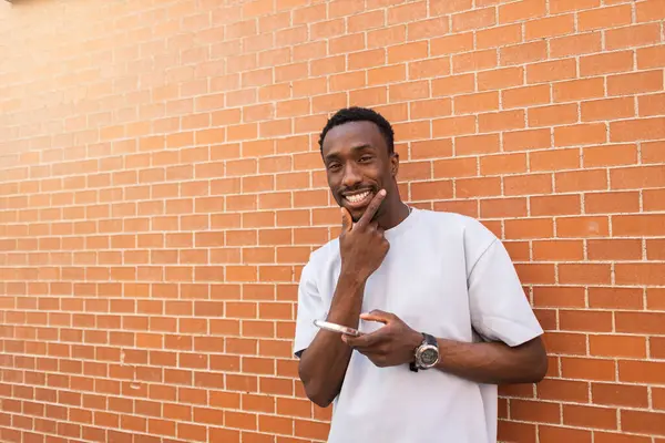 Młody Mężczyzna Trzyma Smartfon Uśmiechając Się Przed Murem — Zdjęcie stockowe