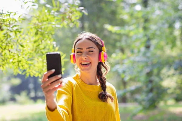 快乐的女人带着耳机在公园里通过智能手机自拍 — 图库照片