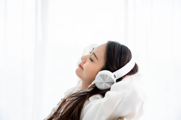 Beautiful young asian woman in headphones listening to music and lying down on bed at bedroom in morning time