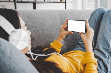 Kanepeye uzanmış, kulaklık takmış, cep telefonu kullanan, beyaz perdeli, beyaz bir kadın. Akıllı telefonu yatay tutuyor. Kırpma yolu dahil olan şablon ekran aygıtı