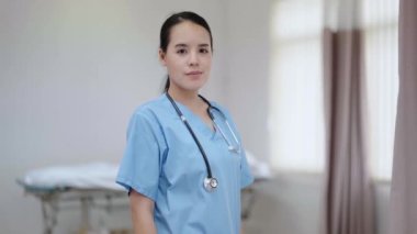Genç, güzel bayan hemşire ayakta kameraya bakarak gülümsüyor.