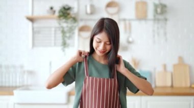 Önlük giyip mutfak odasında duran mutlu Asyalı kadın. Güzel bayan gülümsüyor ve kameraya bakıyor. Kendinden emin hissediyor.