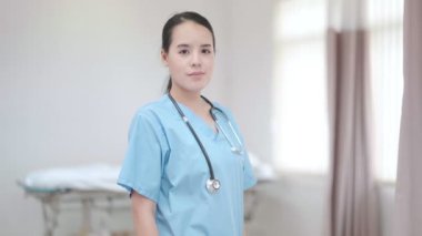 Genç, güzel bayan hemşire ayakta kameraya bakarak gülümsüyor.