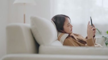 Mutlu genç Asyalı kadın rahat bir kanepede rahat rahat oturup akıllı telefonuna mesaj atıyor, gülümseyen kız cep telefonu kullanıyor, cihaz üzerinde kablosuz internet kullanıyor, evden online alışveriş yapıyor.