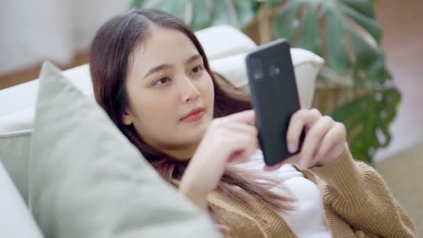 快乐的年轻亚洲女人在家里舒服的沙发上放松 用智能手机发短信 微笑的女孩用手机 在电子产品上浏览无线网络 从家里上网购物 — 图库视频影像