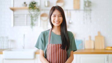 Önlük giyip mutfak odasında duran mutlu Asyalı kadın. Güzel bayan gülümsüyor ve kameraya bakıyor. Kendinden emin hissediyor.