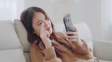 Happy young asian woman relax on comfortable couch at home texting messaging on smartphone, smiling girl use cellphone, browse wireless internet on gadget, shopping online from home