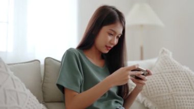Mutlu genç Asyalı kadın evdeki rahat kanepede dinleniyor, akıllı telefondan online oyun oynuyor, gülümseyen kız cep telefonu sohbetini kullanıyor, cihazda kablosuz interneti tarıyor, evden online alışveriş yapıyor.