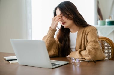 Ofis sendromu. Çok çalışan yorgun Asyalı kadın baş ağrısı çekiyor, bilgisayardan sonra görme sorunu yaşıyor. Stresli genç kadın evde yorgunluk çekiyor.