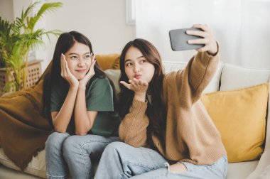 İki Asyalı genç kadın mutlu mesut gülümsüyor ve evdeki oturma odasında selfie çekiyor.