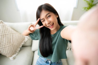 Oturma odasında oturan kocaman gülüşlü Asyalı güzel bir bayan. Bulanık arka planda neşeli selfie çekerken eğleniyor.