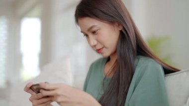 Mutlu genç Asyalı kadın evdeki rahat kanepede dinleniyor, akıllı telefondan online oyun oynuyor, gülümseyen kız cep telefonu sohbetini kullanıyor, cihazda kablosuz interneti tarıyor, evden online alışveriş yapıyor.