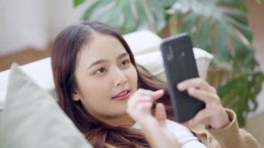 Mutlu genç Asyalı kadın rahat bir kanepede rahat rahat oturup akıllı telefonuna mesaj atıyor, gülümseyen kız cep telefonu kullanıyor, cihaz üzerinde kablosuz internet kullanıyor, evden online alışveriş yapıyor.