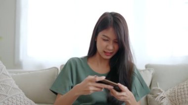 Mutlu genç Asyalı kadın evdeki rahat kanepede dinleniyor, akıllı telefondan online oyun oynuyor, gülümseyen kız cep telefonu sohbetini kullanıyor, cihazda kablosuz interneti tarıyor, evden online alışveriş yapıyor.