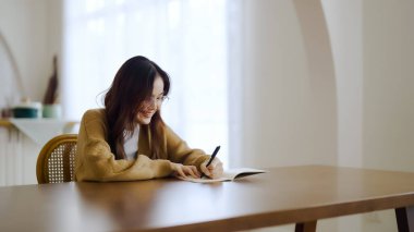 Güzel genç Asyalı kadın evde oturuyor ve not alıyor. Çevrimiçi çalışırken mutlu gülümseme, evde serbest çalışma