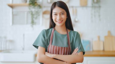 Önlük giyip mutfak odasında duran mutlu Asyalı kadın. Güzel bayan gülümsüyor ve kameraya bakıyor. Kendinden emin hissediyor.