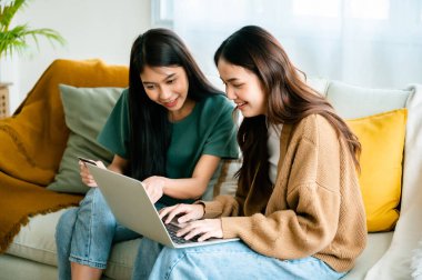 Two asian young woman happy smiling and using computer laptop on couch in living room at home