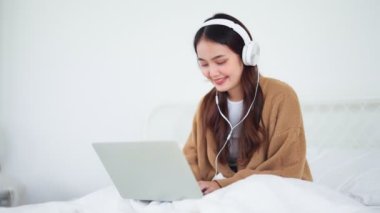 Happy beautiful young asian woman working on computer laptop and sitting on bed in house. She making video conference webinar and wearing headphone listening music