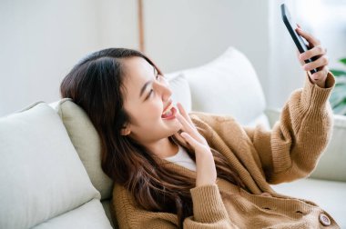 Mutlu Asyalı kadın evde dinleniyor. Kadın kanepede oturuyor ve cep telefonuyla video görüşmesi yapıyor.