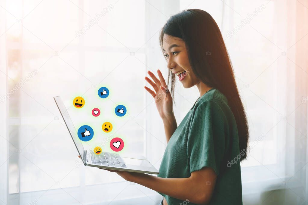 Young asian woman happy smiling and using computer laptop with icon social media and social network beside window