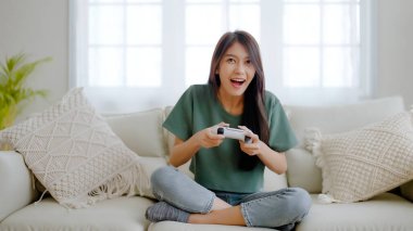 Güzel, genç Asyalı bir kadın kanepede oturuyor ve elinde joystick ile evdeki oturma odasında oyun oynuyor.