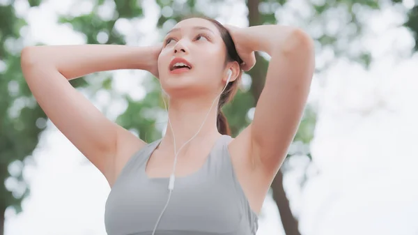 Asian woman trainer is engaged in fitness in public park. Female wearing sport bra and using headphone listening to music. Healthy lifestyle, sports outdoor activities in park