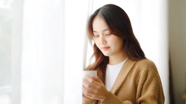 Genç Asyalı kadın pencerenin yanında duruyor ve elinde kupayla oturma odasında sabah uyandıktan sonra süt içiyor.