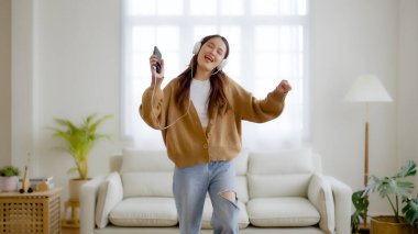 Evdeki oturma odasında, kulaklık takıp yerde dans eden Asyalı genç bir kadın. Mutlu Asyalı kadın gülümsemesi evde dinleniyor, sağlıklı zihinsel sağlık ve refah.