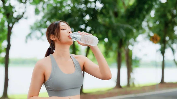 Young asian woman drinking water from a bottle while taking a break during her workout. Happy adolescence female working out in the park and taking rest a break to drink water