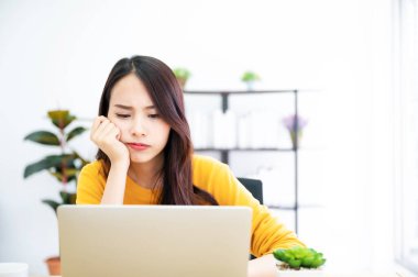 Asyalı genç bir kadın ciddi ciddi evdeki bilgisayar dizüstü bilgisayarında çalışıyor. Çözüm bulma sorununu düşünüyor.