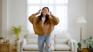 Evdeki oturma odasında, kulaklık takıp yerde dans eden Asyalı genç bir kadın. Mutlu Asyalı kadın gülümsemesi evde dinleniyor, sağlıklı zihinsel sağlık ve refah.