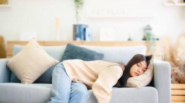 Asian woman resting at home on couch, feeling exhausted after work, lacking energy, or overworked, too tired, and lacking motivation clipart