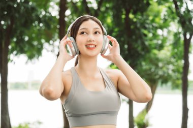 Asian woman trainer is engaged in fitness in public park. Female wearing sport bra and using headphone listening to music. Healthy lifestyle, sports outdoor activities in park