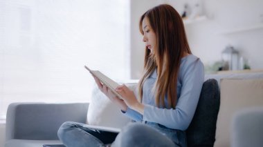 Genç Asyalı kadın evdeki kanepede otururken kitap okuyor.