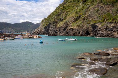 Vernazza, Cinque Terre, Italy, Liguria - May 12, 2019:  View of the Mediterranean Sea coast, turquoise water with moored boats and surrounding rocks, stone pier with walking tourists