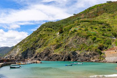 Vernazza, Cinque Terre, Italy, Liguria - May 12, 2019:  View of the Mediterranean Sea coast, turquoise water with moored boats and surrounding rocks, stone pier with walking tourists