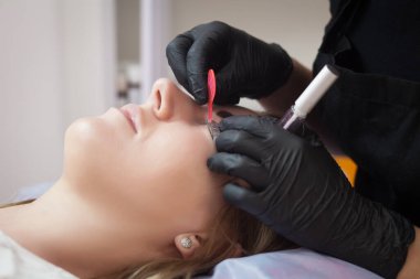 Güzel genç bir kadın kaş düzeltmesi yaptırıyor ve göz kapaklarını kapatıyorlar. Güzellik salonunda genç bir kadın gözlerini boyuyor..