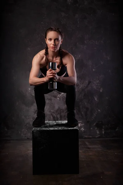 Beyaz bir kız dambıllarla meşgul. Küpün üstüne çömel. Stüdyo arkaplanı