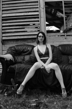 Koyu renk iç çamaşırlı güzel esmer kadın, tahta evin yanındaki deri koltukta poz veriyor. 