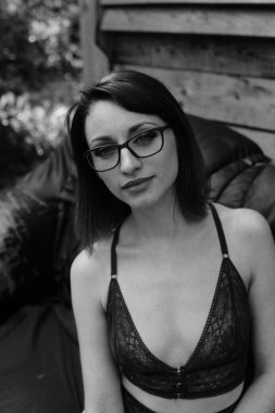 Koyu renk iç çamaşırlı güzel esmer kadın, tahta evin yanındaki deri koltukta poz veriyor. 