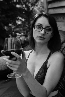 Koyu renk iç çamaşırı giymiş, elinde şarap kadehi olan ve dışarıdaki ahşap evin yanındaki deri koltukta poz veren güzel esmer kadın. 
