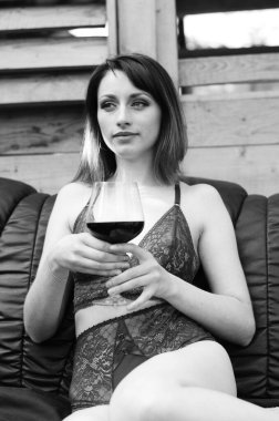 Koyu renk iç çamaşırı giymiş, elinde şarap kadehi olan ve dışarıdaki ahşap evin yanındaki deri koltukta poz veren güzel esmer kadın. 